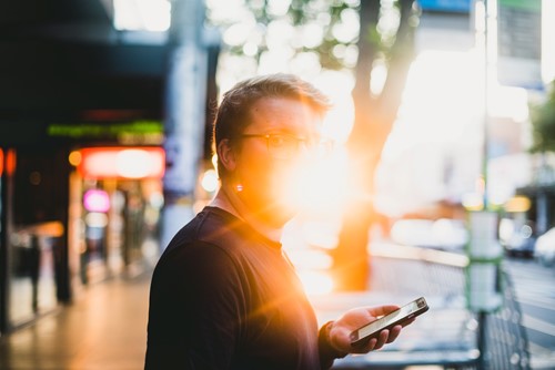 A man stands on a busy street holding his phone backlit by bright sunlight, masking his face.